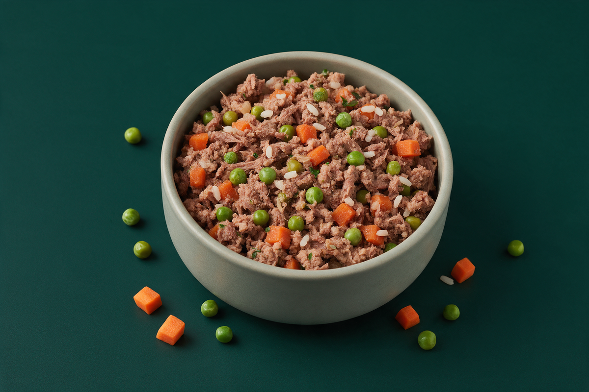 A full bowl of gently cooked dog food on a deep green background with a few loose peas and carrot dice scattered beside it
